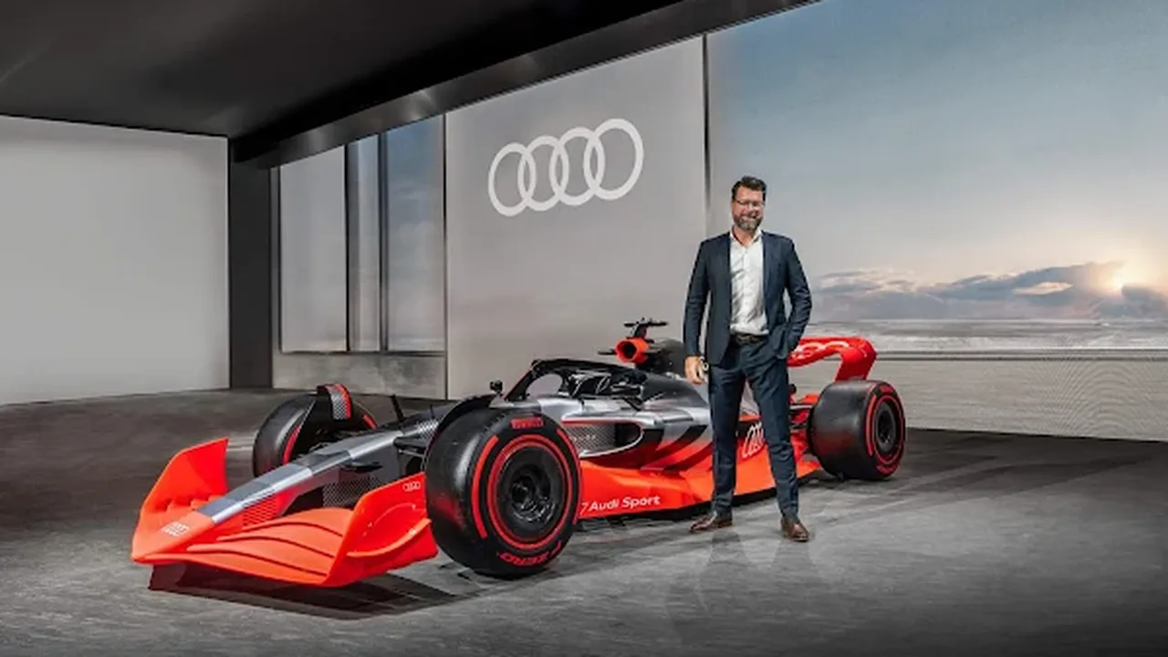 Logo da Audi sobre fundo do paddock da Fórmula 1 com carros da Sauber ao fundo